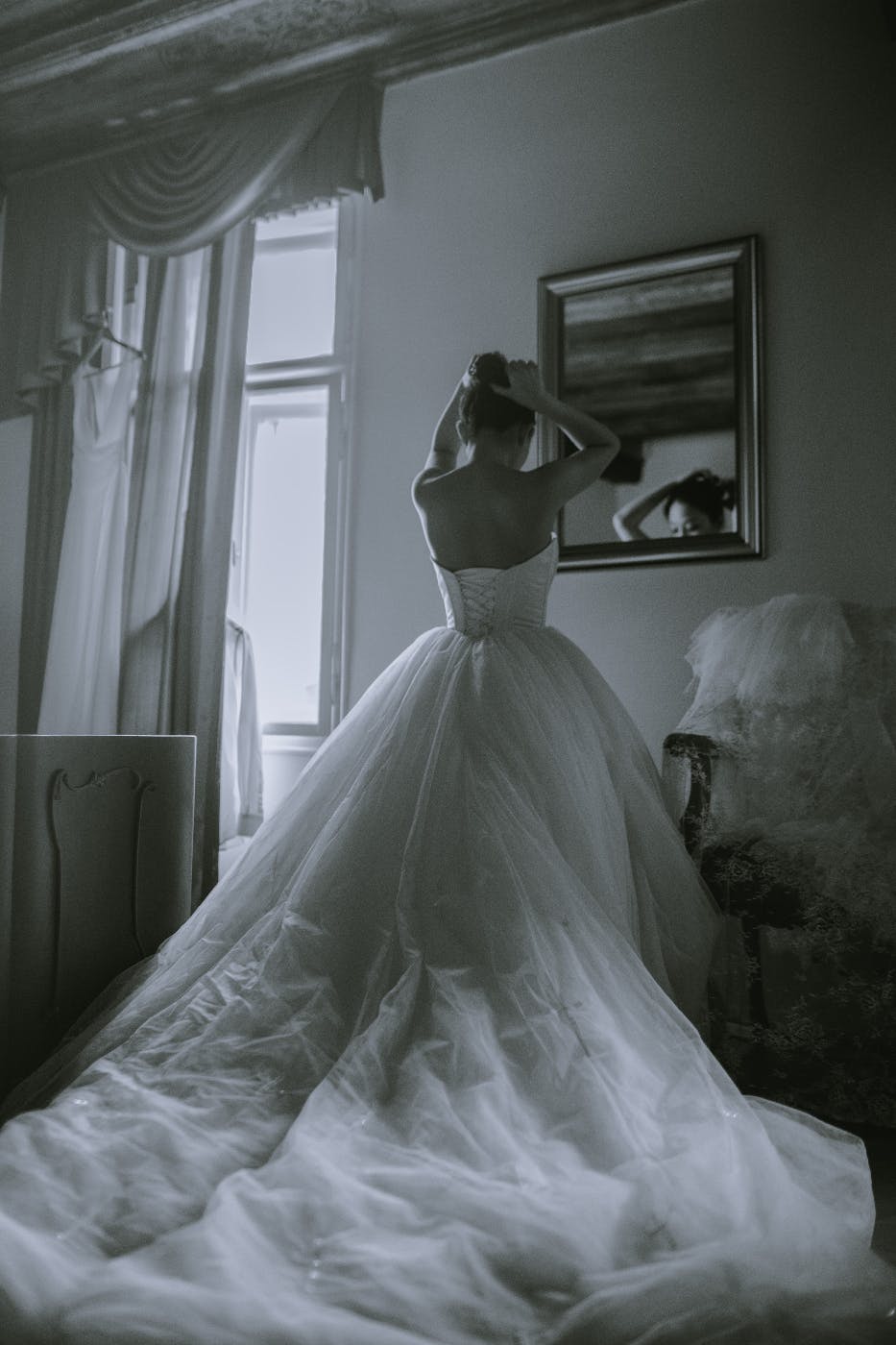 A black and white photo of a bride in her wedding dress fixing her hair in a mirror