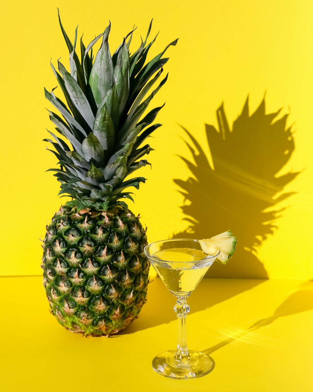 a pineapple next to a martini with a yello bakcground and the shadow of the pineapple ont he wall