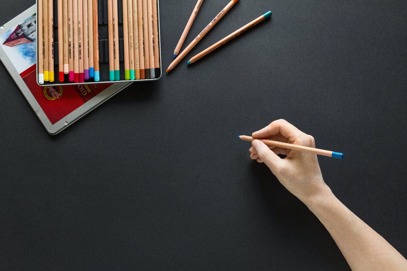 A tin of colored pencils and a hand holding one about to draw on a black surface