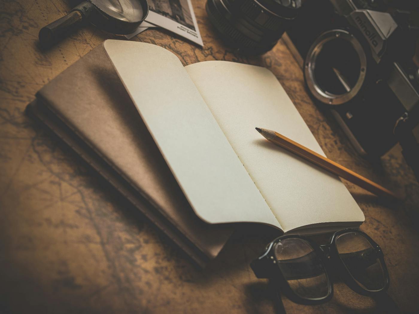 Open journal on a table with a camera, a pair fo glasses, and a pencil.