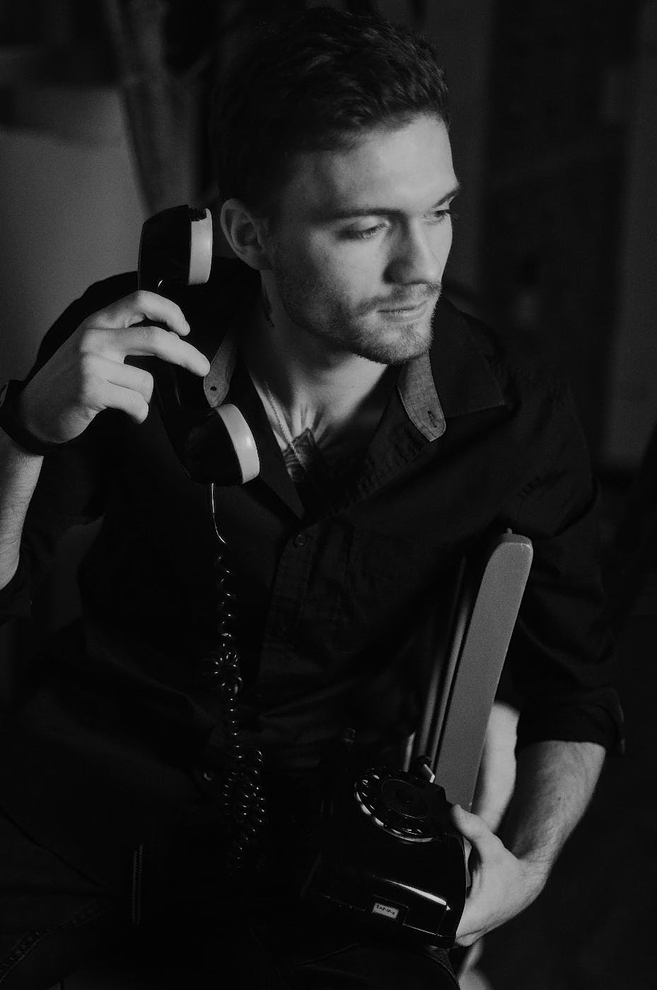Black and white photo of a young man sitting in a chair using a rotary phone