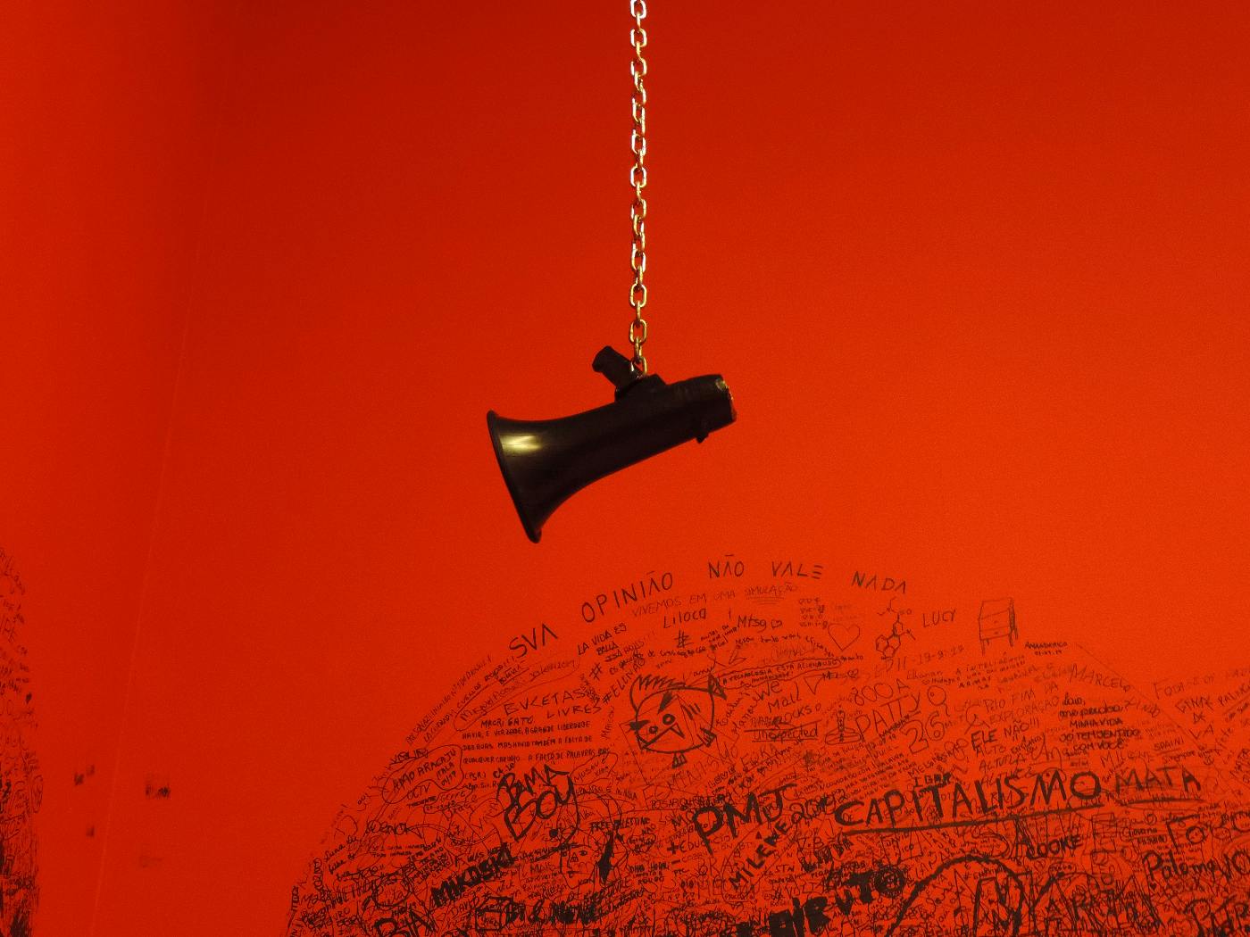 A magaphone hanging at the end of a chain against a red graffitied wall