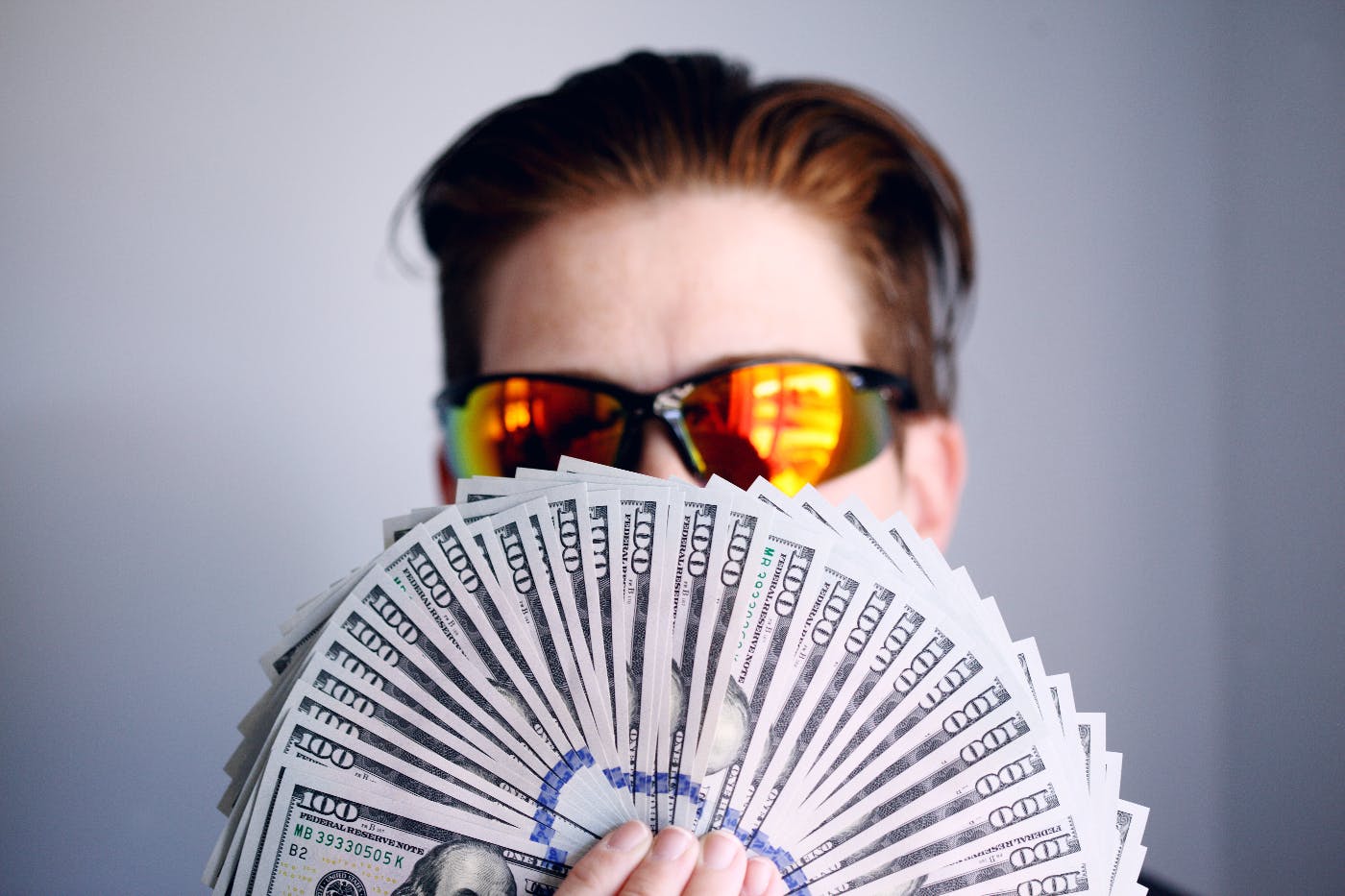 A man wearing reflective yellow sunglasses holding a fan of 100 dollar bills