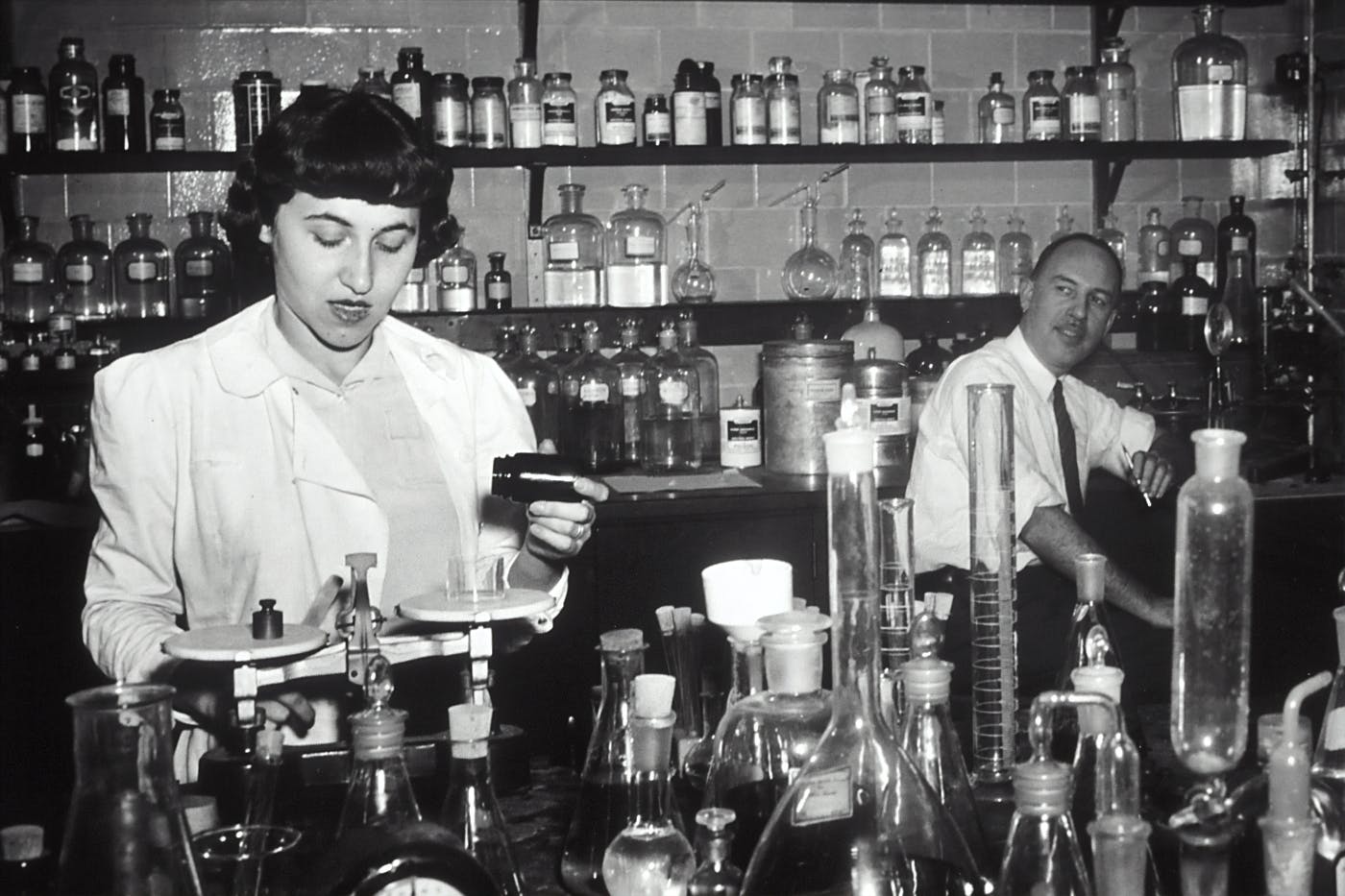 a black and white photo of a male and female scientists working with tubes, pills and bottles.
