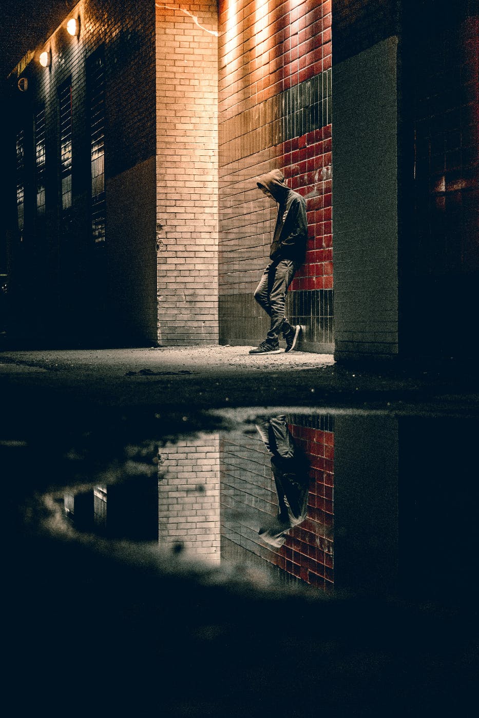 A man leaning against a red tile wall, night. he is reflected in a puddle