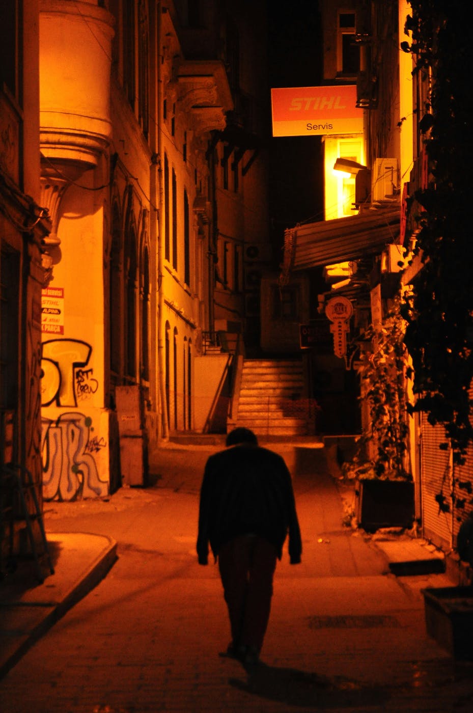 A man walking an empty side street at night lit by amber neon light