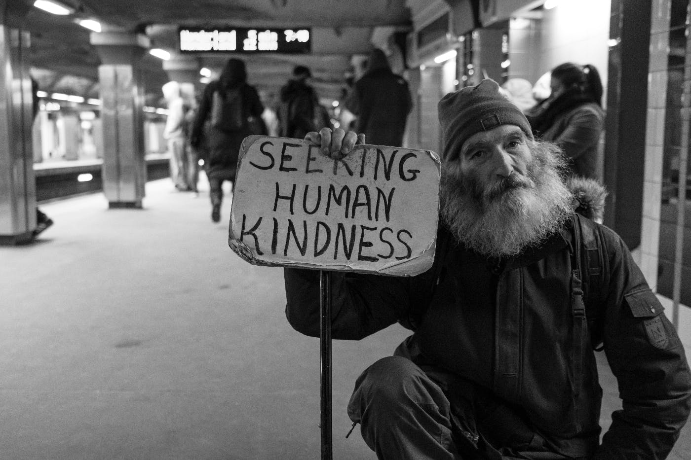 a homeless man in a subway station holding a sign that reads: Seeking Human Kindness