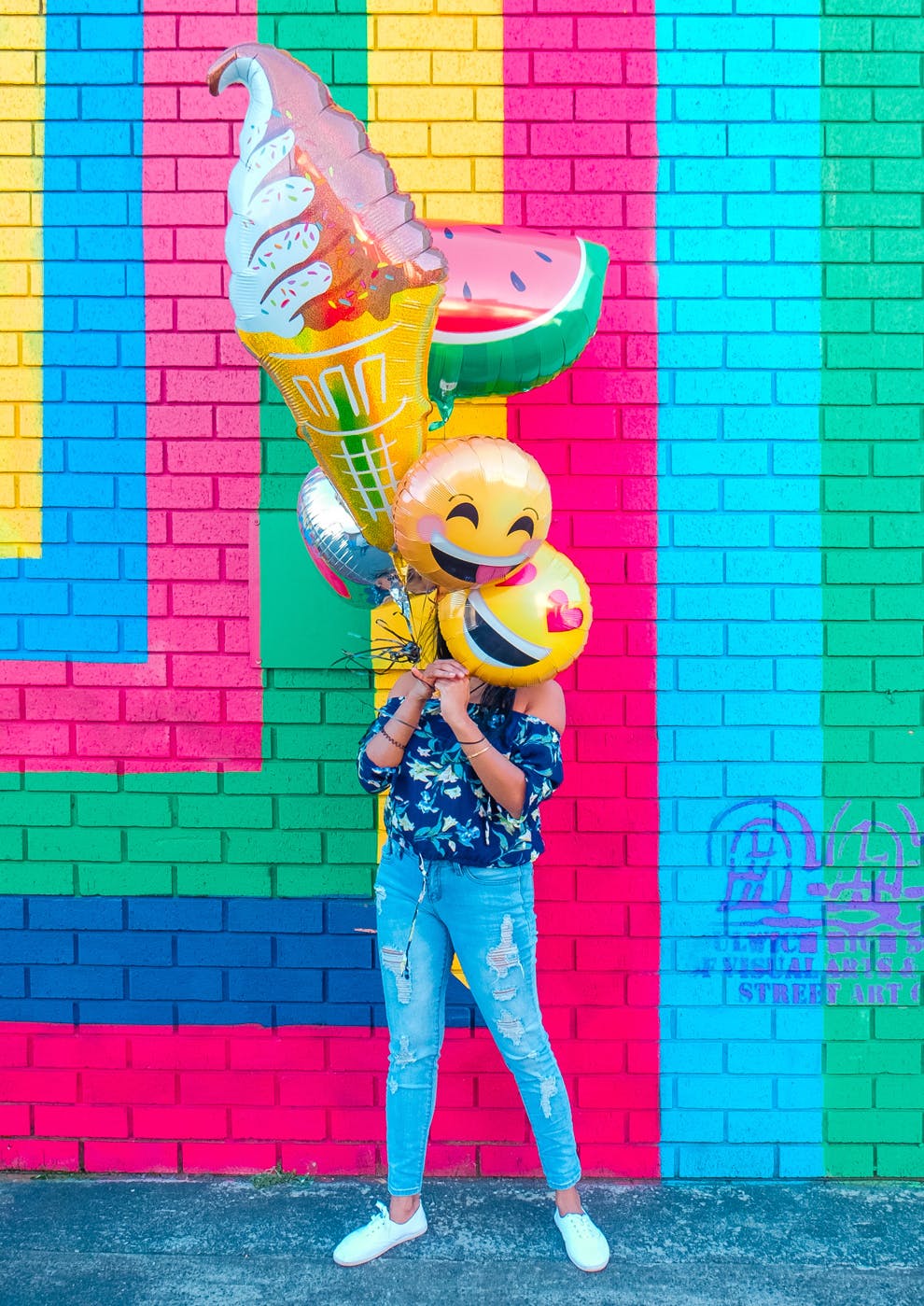 woman standing before a colorful wall hiding her face behind large fruit shaped balloons