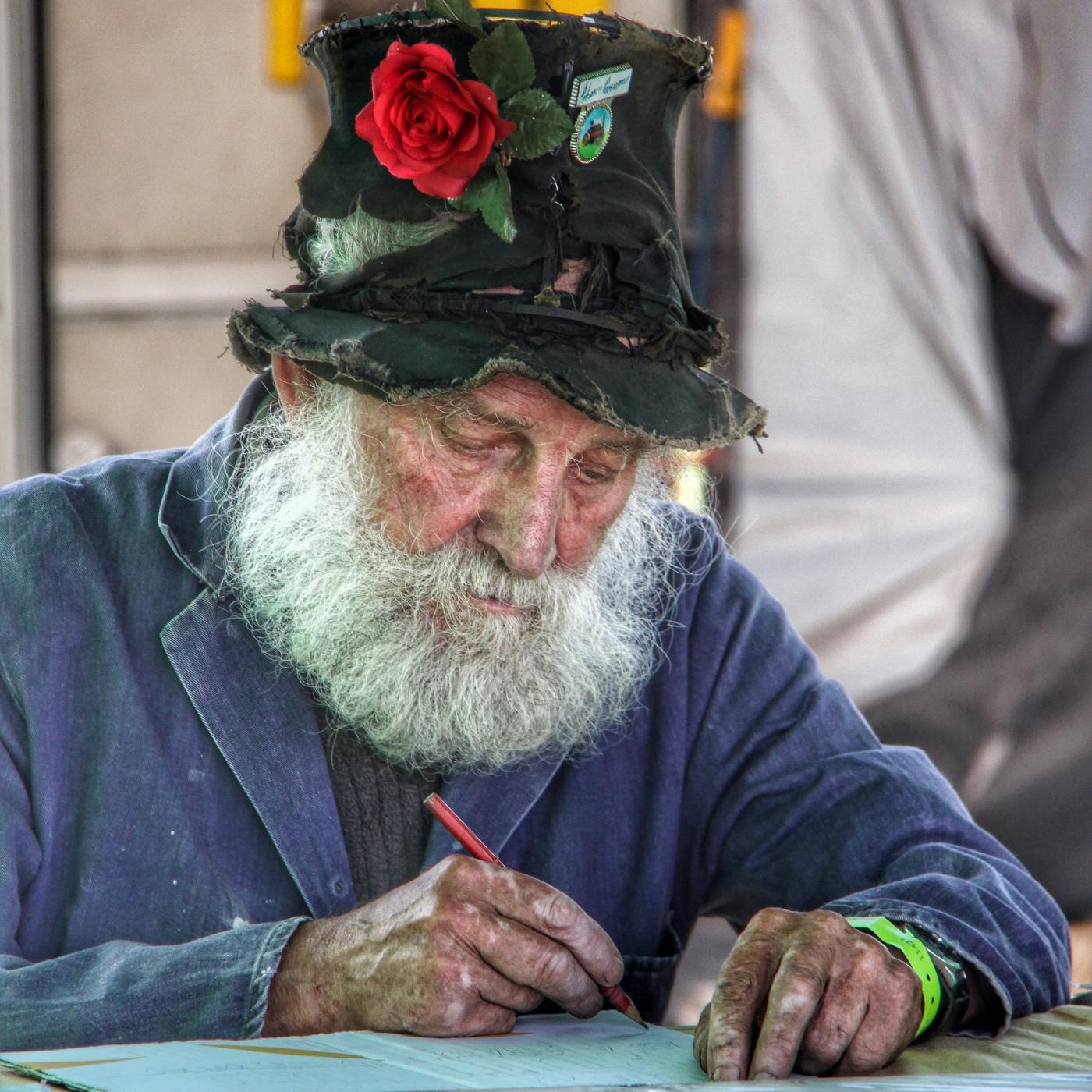 An older man in a ragged, flowered hat writing at a table with a red pencil