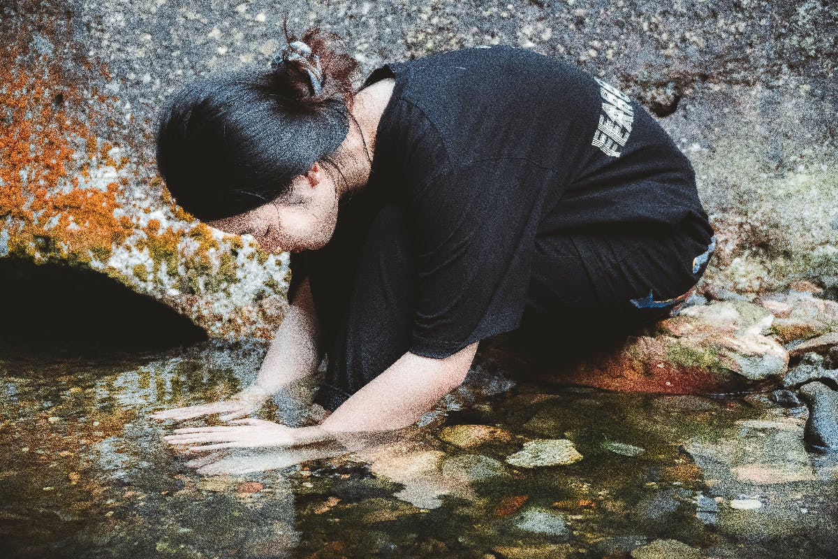 awoman kneeling by a spring with her hand sin the water to her elbows, eyes closed meditating