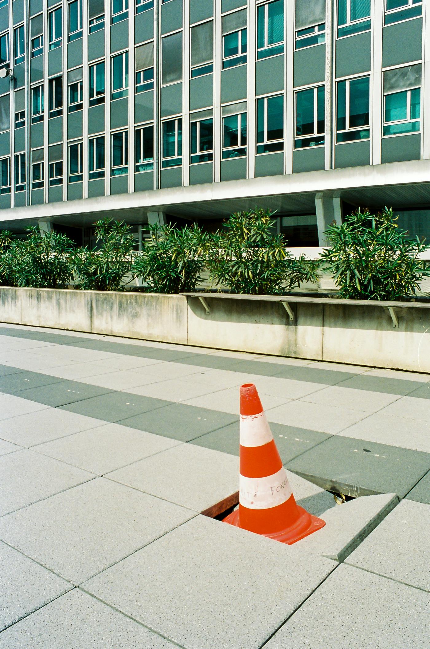 An orange and white traffic cone sitting on a broken croncrete sidewalk tile