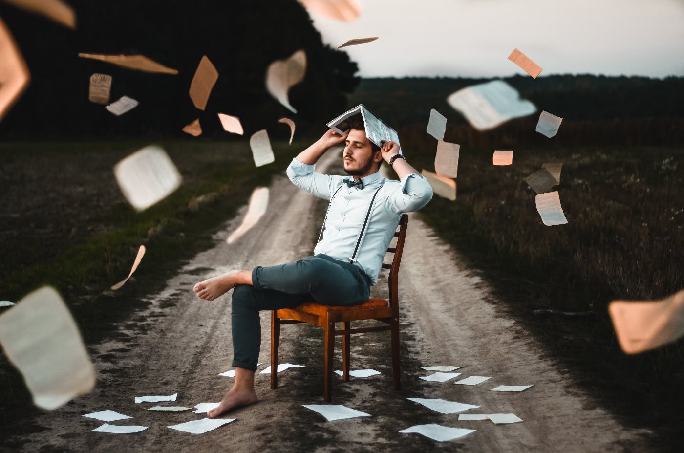 A barefoot man seated in a chair with an open book on his head surrounded by flying papers