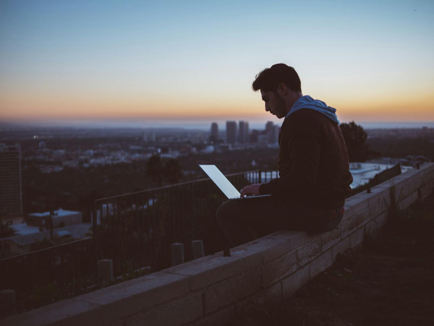 A guy sitting on the edge of a wall with a cityscape behind him working on a laptop.