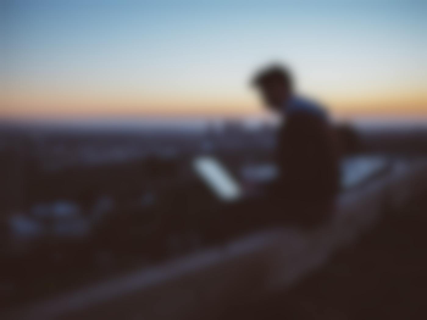 A guy sitting on the edge of a wall with a cityscape behind him working on a laptop.