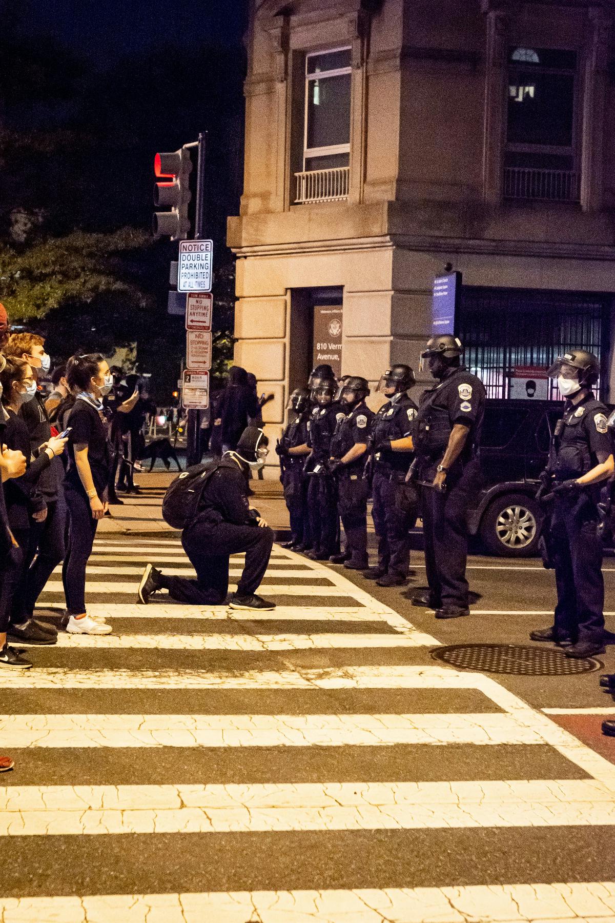 Protesters facing a police line with one protester kneeling for peace