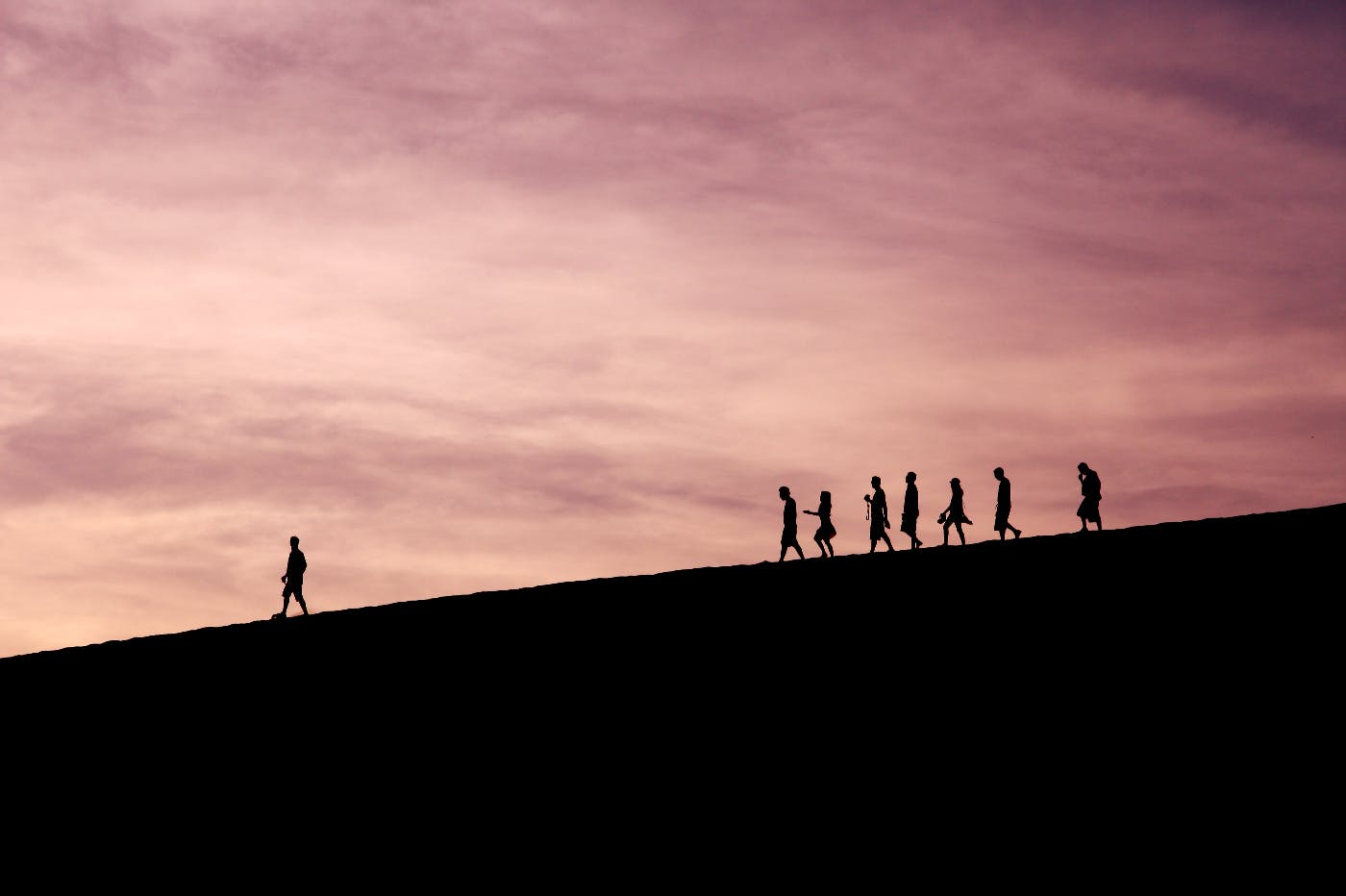 Silhouette of people walking down a hill with one person far in front of the otehrs leading