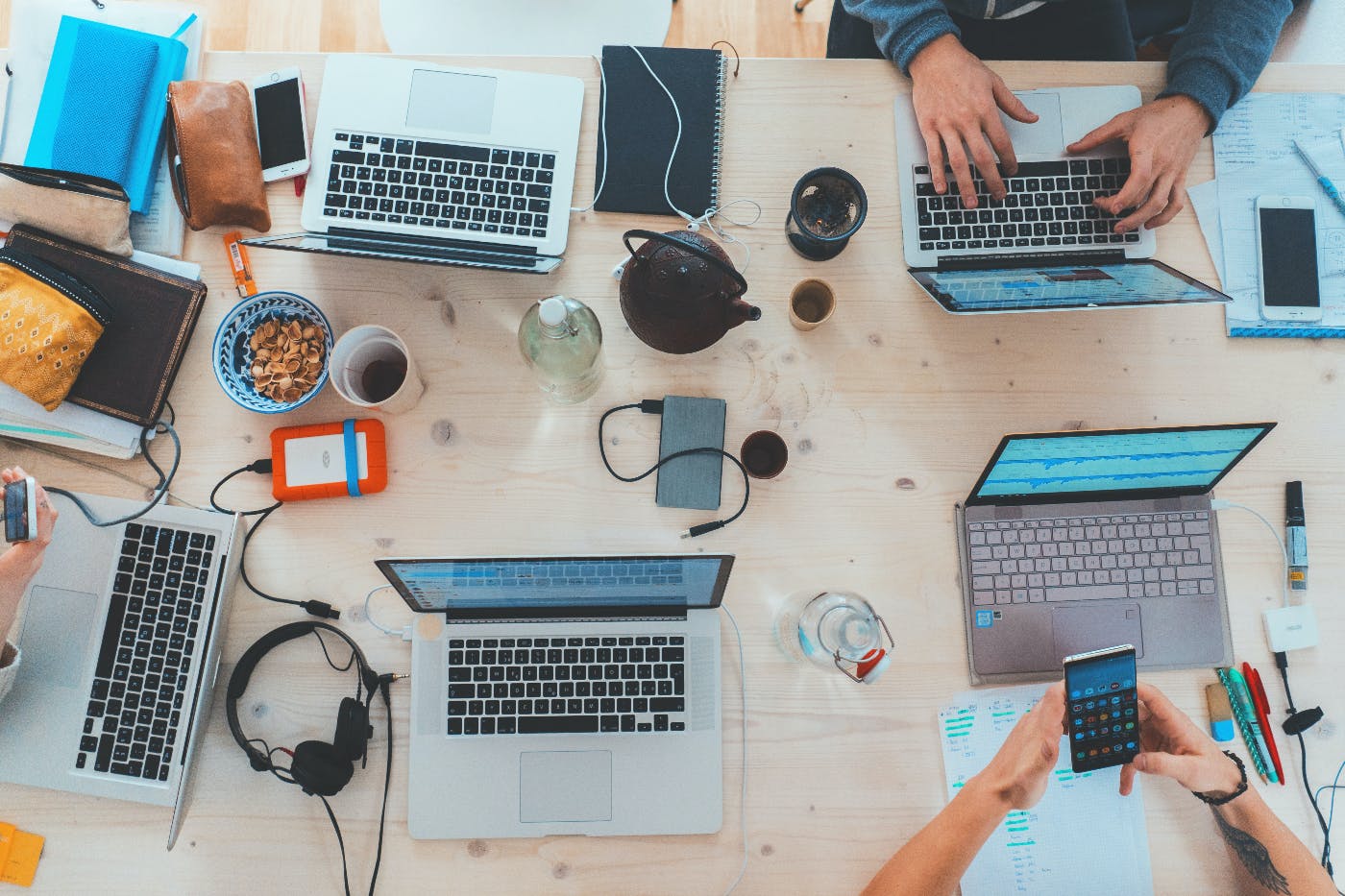 A table with 6 laptops, 6 smartphines with headsets, coffee cups m snakc bowls and two people working.