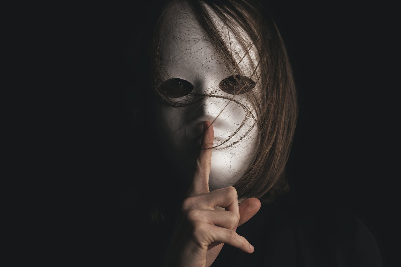 a person with long hair, wearing a white mask with their finger held to their lips in a shushing motion