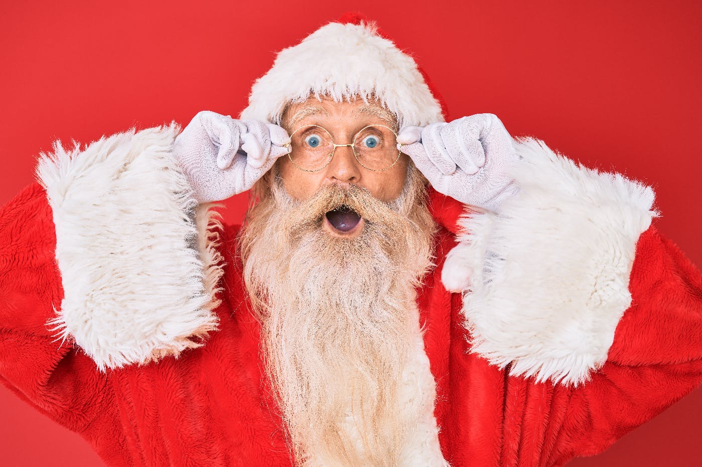 A man dressed as Santa with a natural beard, holding his round glasses and giving a open mouth smile
