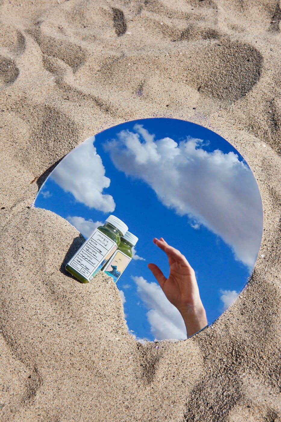 a round mirror in the sand with a small vile and clouds and a hand reflected