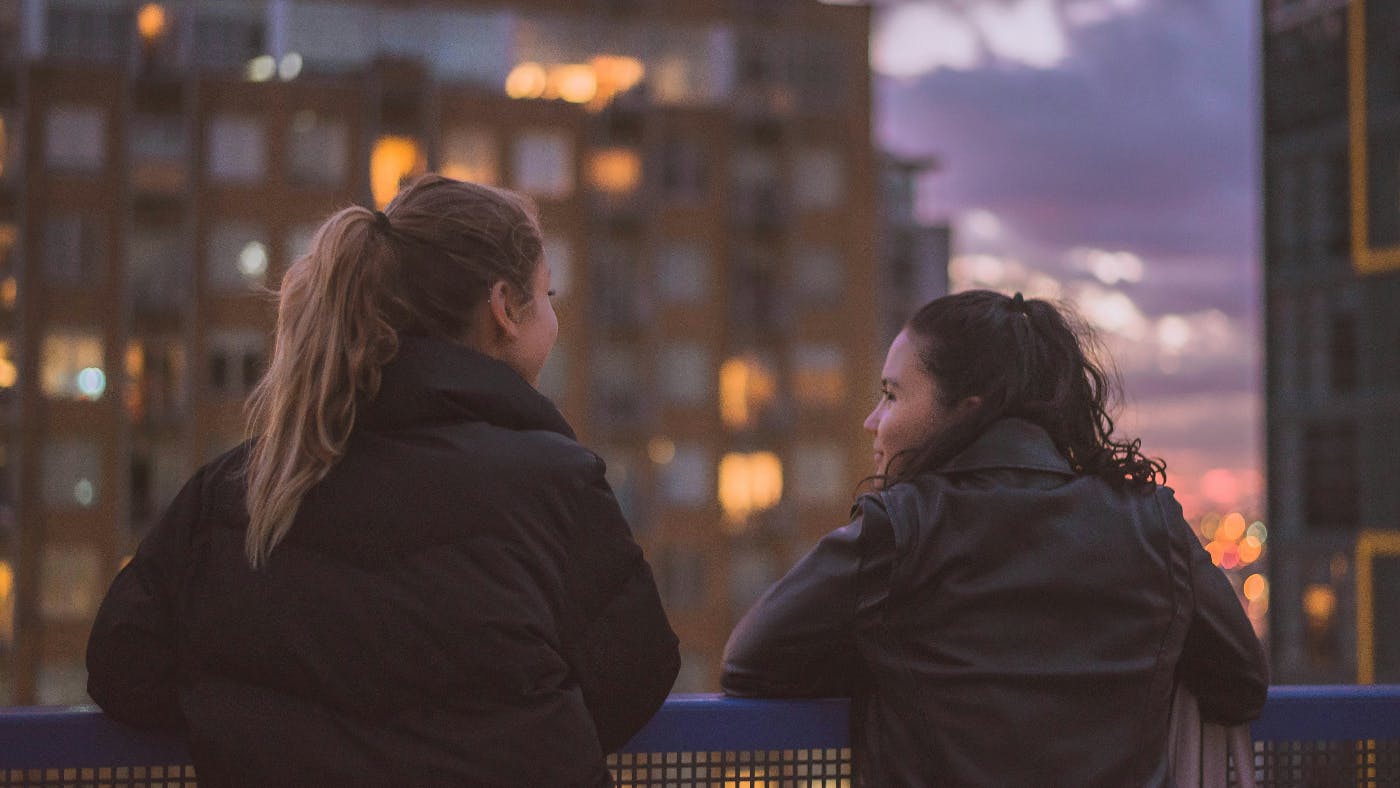 Two girls standing on a balcony overlooking a city having a conversation