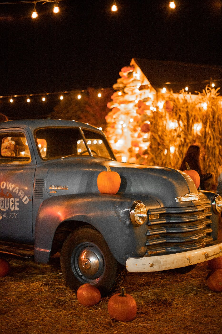 an old pickup truck with pumpkins on the fenders in front of corn stalks and hanging lights