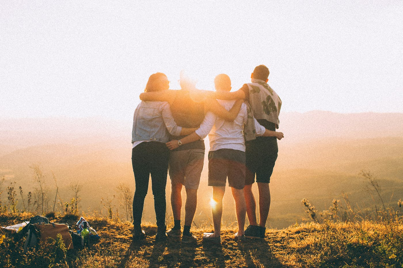 four people. backs to the camera, standing on a hill, watching the sun set with their arms around each other