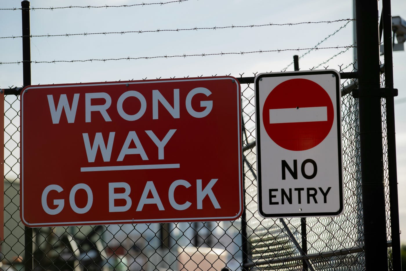 Two signs on a chain link fence topped with barbed wire: Wrong Way Go Back & No Entry