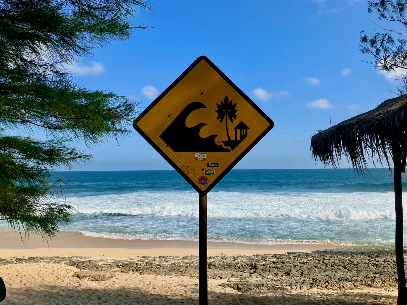 A yellow sign on a beach depicting a giant wave encroaching on a palm tree and a cottage