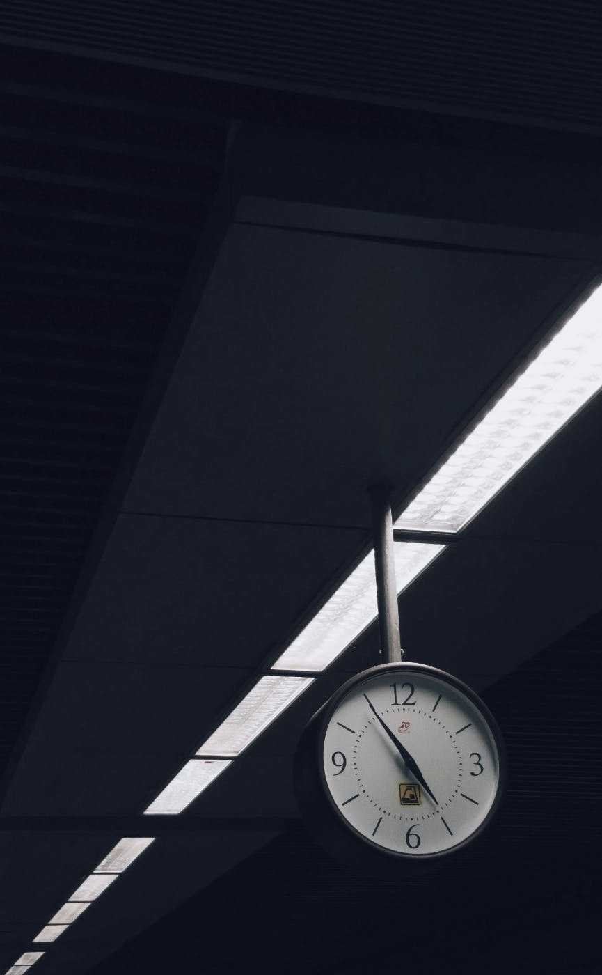 A clock hanging from a ceiling by some fluorescent lights it is 5 til 5