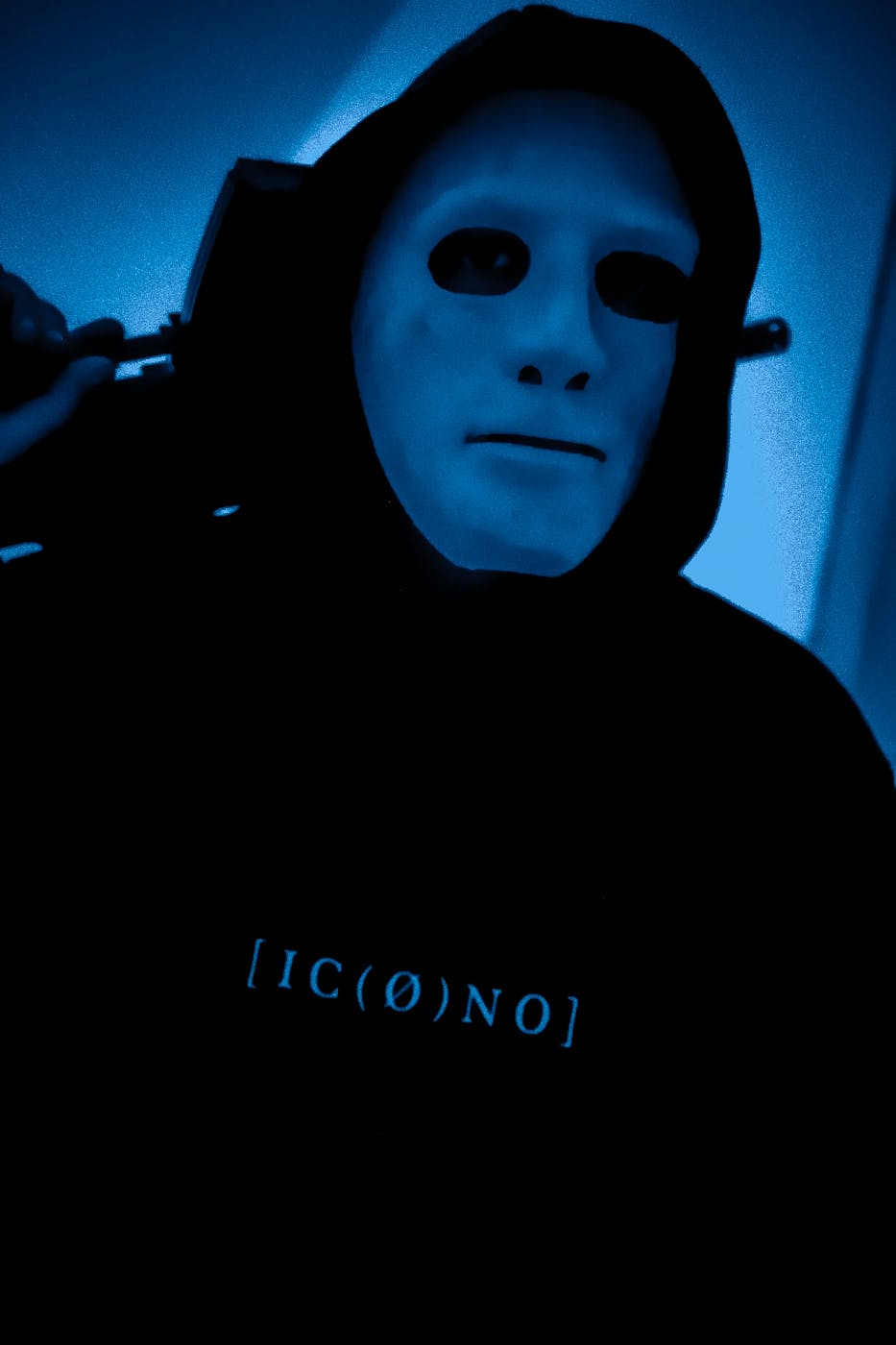 A man in a black hoodie wearing a white human mask under blue light