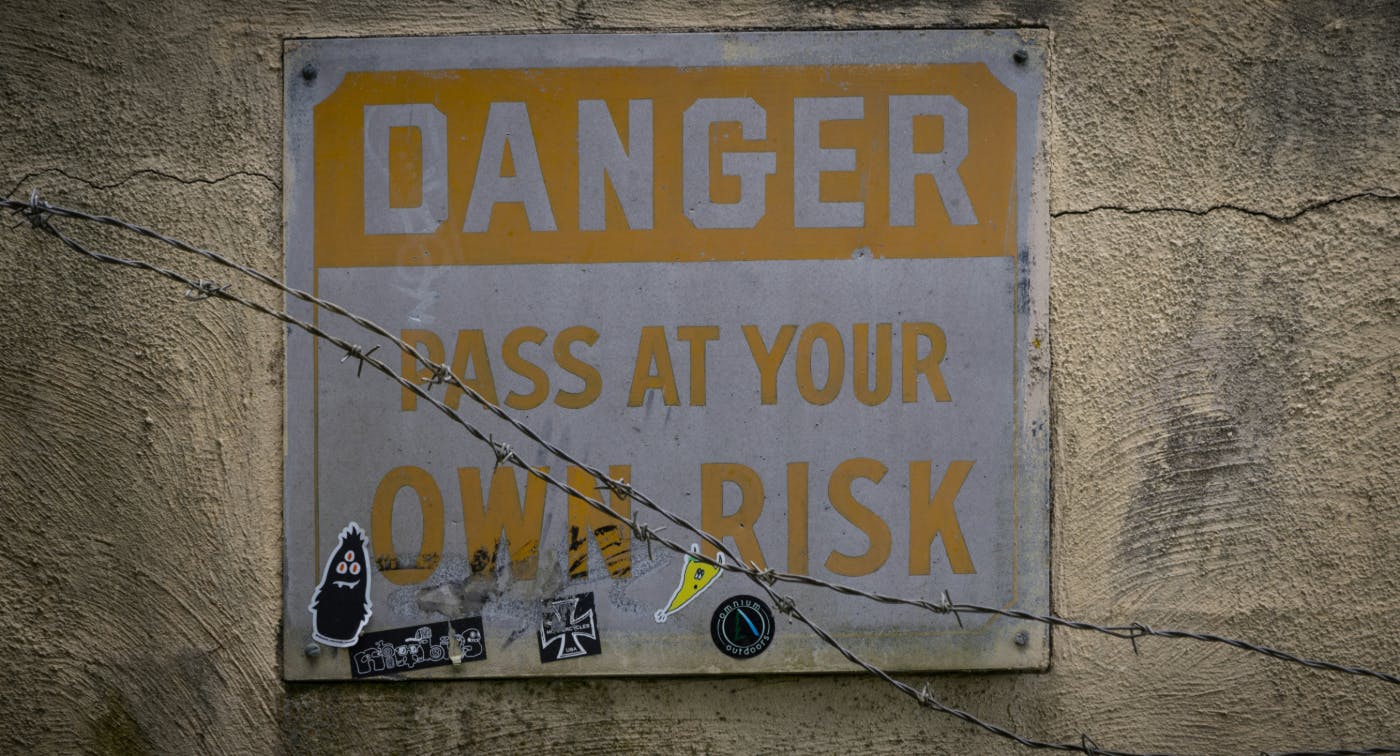  yellow and white sign reading Danger pass at your own risk on a wall behind some barbed wire