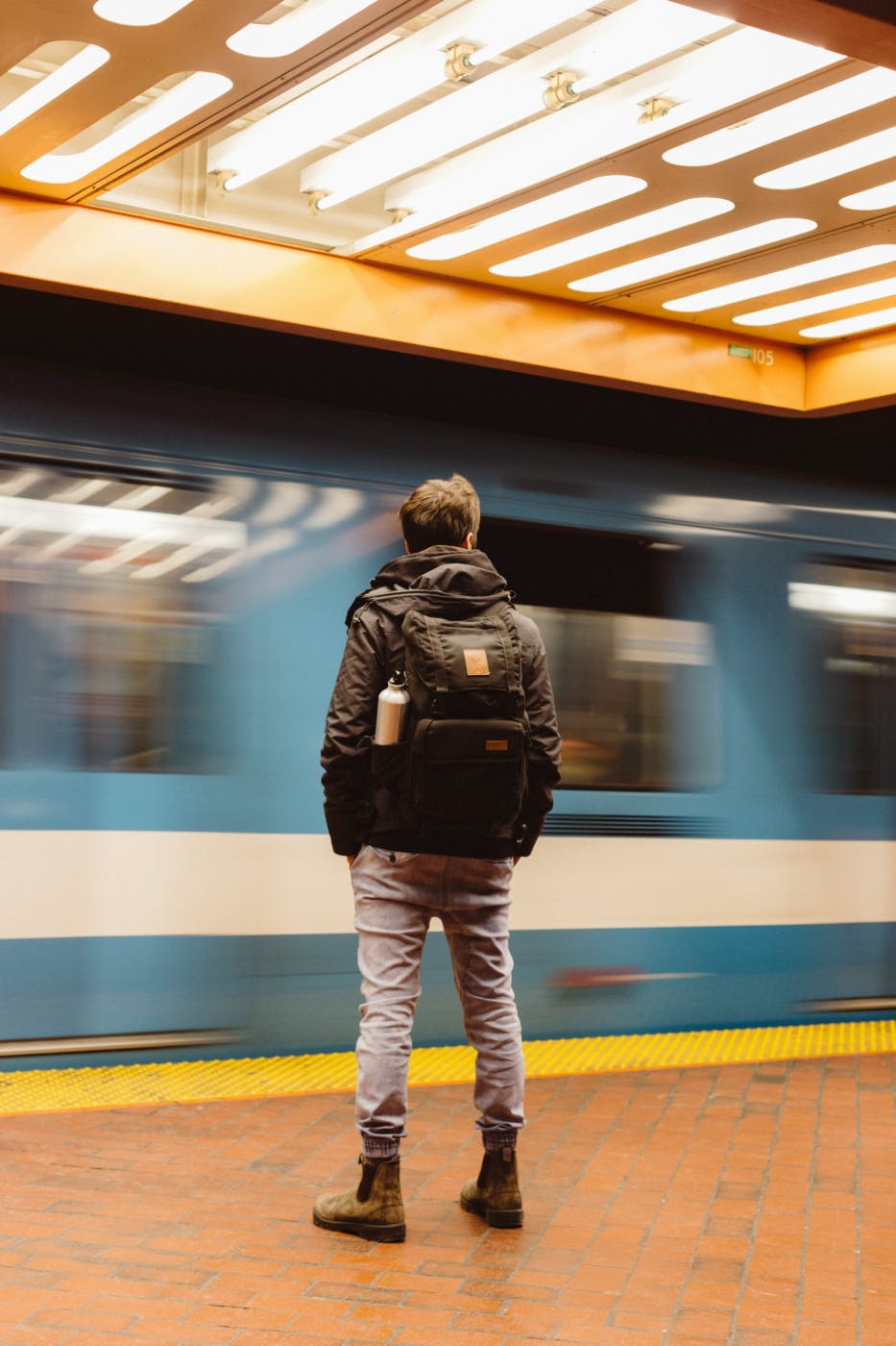  guy with a backpack standing on a subway platform and a train races by