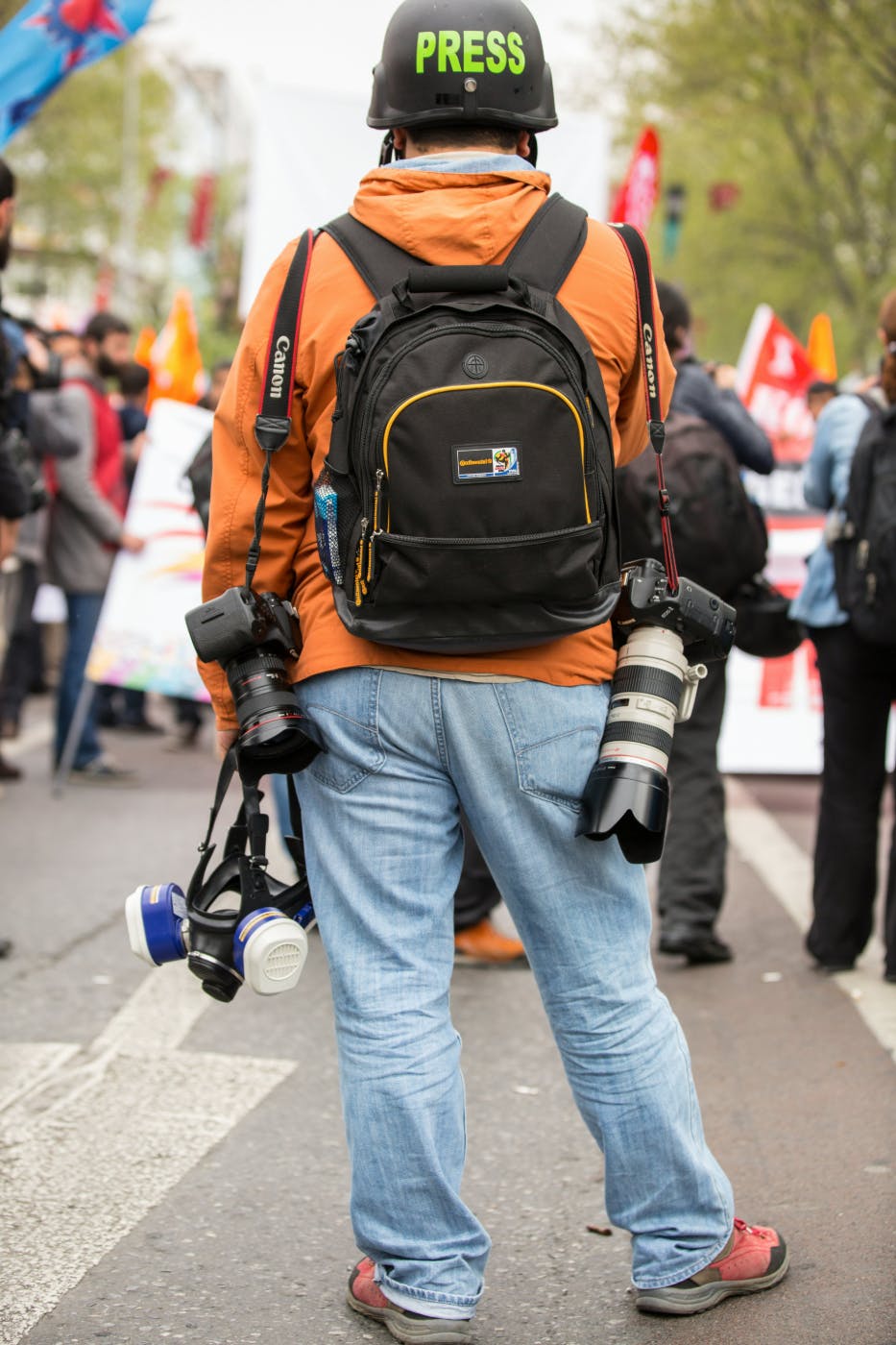 A man in an orange hoodie, carrying 2 cameras and a gas mask, wearing a helmet with PRESS written on it.