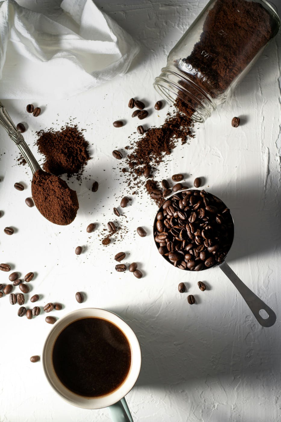 A white table cloth with, a jar of ground coffee. a scoop of coffee beans, a cup of coffee and a spoon of ground beans