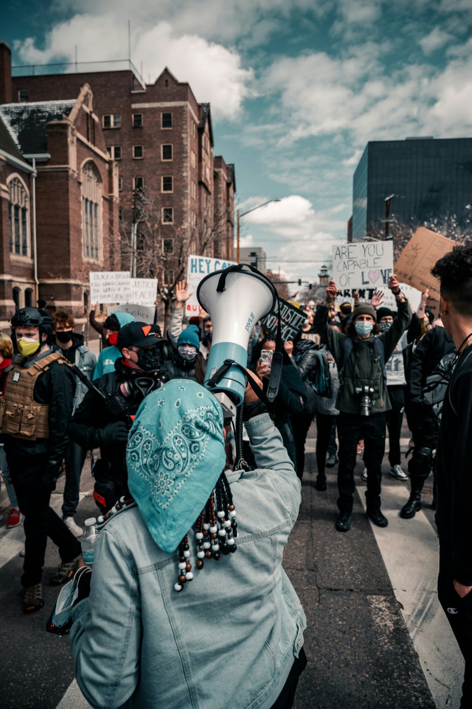 A girl with beaded hair holding a megaphone addressing a crowded street in protest