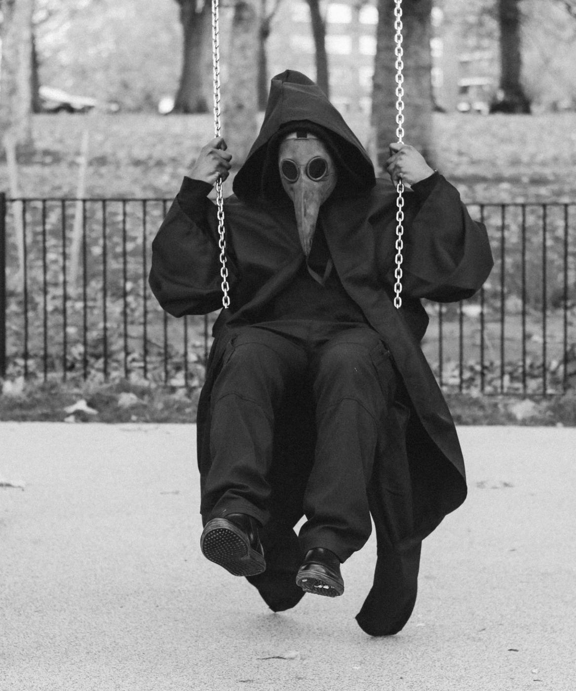 A man all in black, wearing a hood and a plague mask on a swing