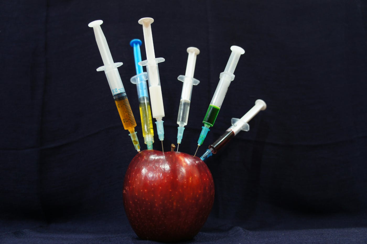 Six syringes with varying colored liquids stuck in a red apple