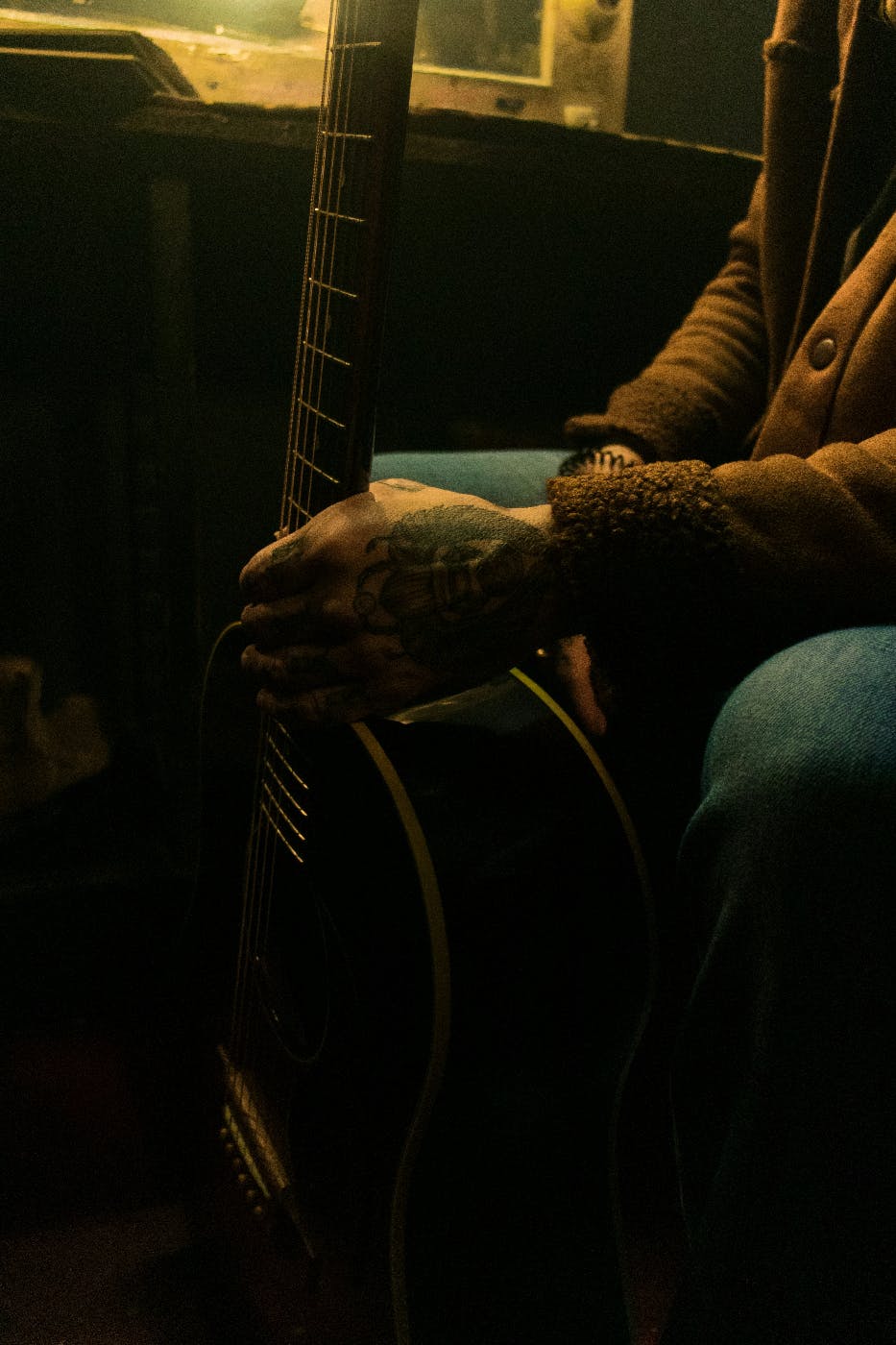 A man in a dimly lit room holding a guitar with a tattooed hand