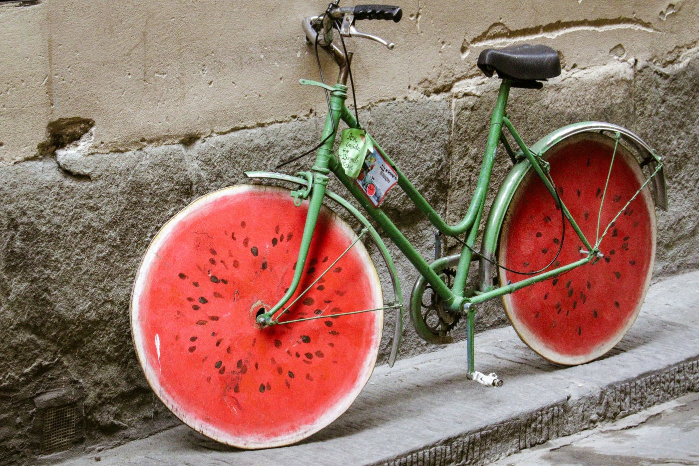 A green bike with wheels that are made to look like watermelon slices leaning against a stone wall