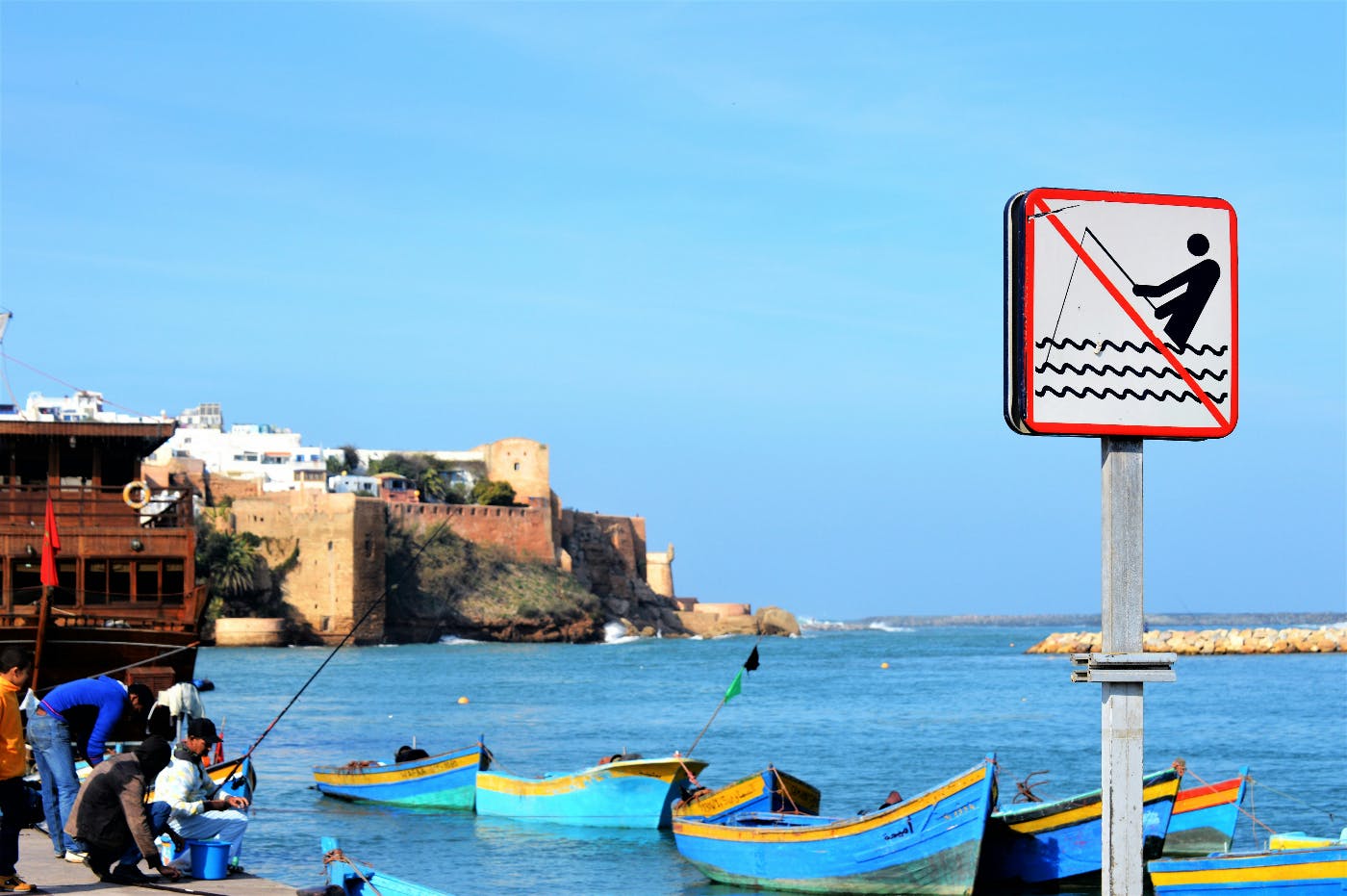 A waterfront with blue boats and several men fishing near a sign with the image of a guy fishing with a line through it.