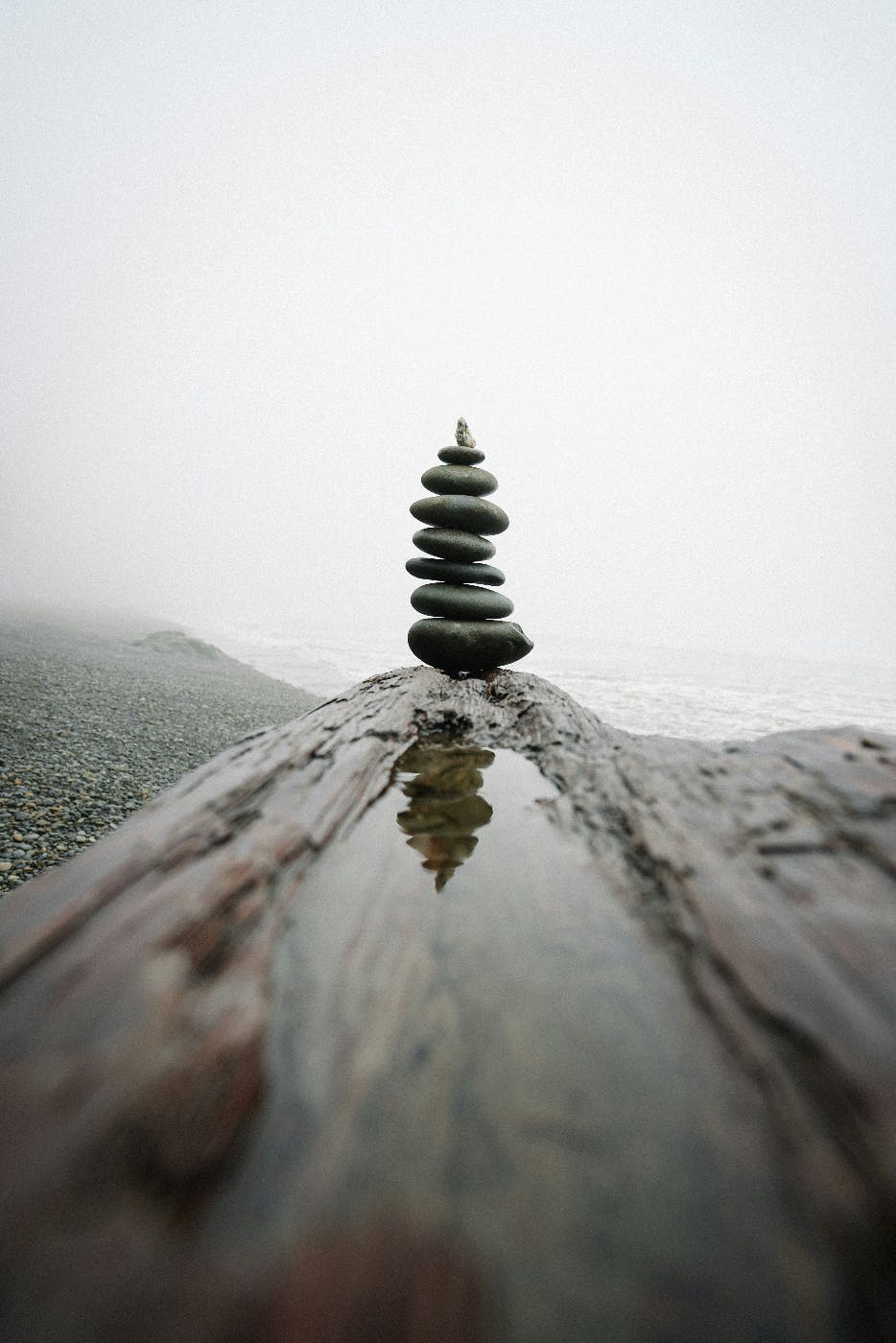 8 stones balanced on each other on a log by the sea, their reflection seen in a puddle on the log