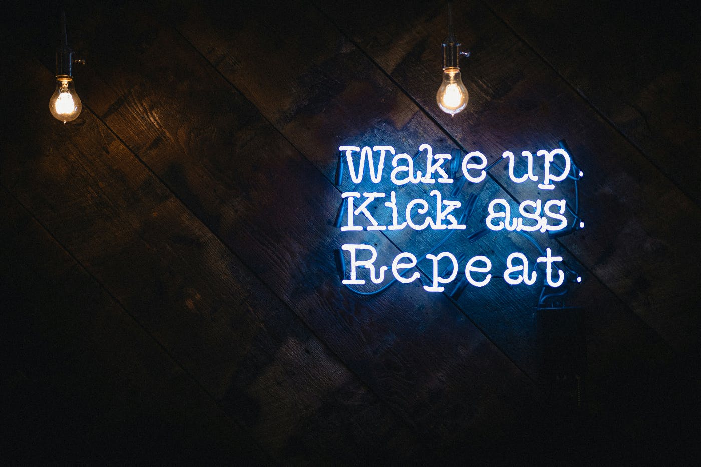 A blue neon sign on a wood wall reading Wake up. Kick ass. Repeat.