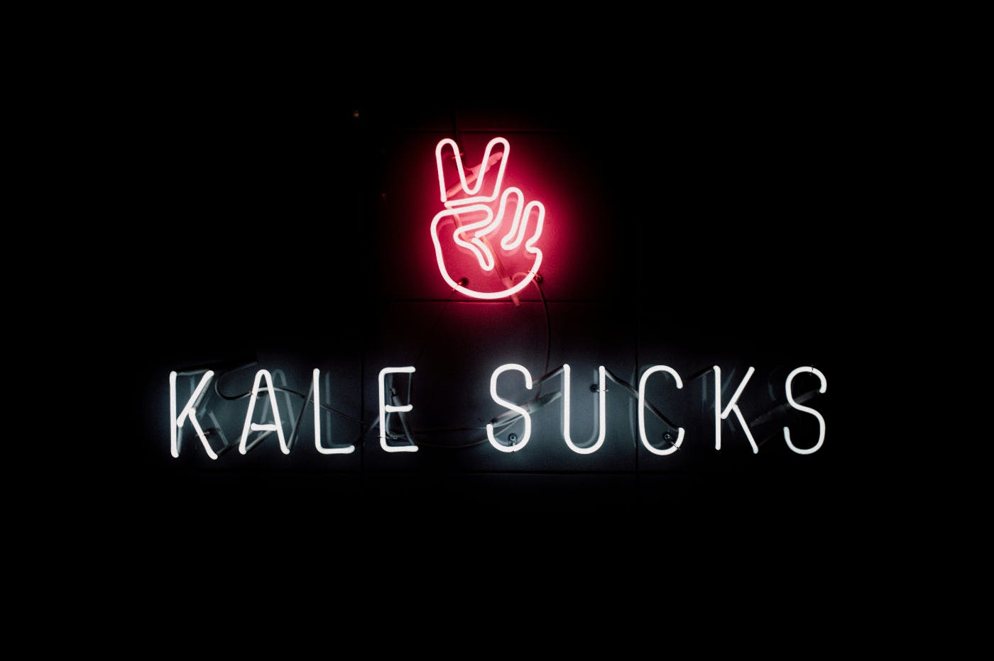 A neon sign, red hand throwing a peace sign, white letters reading Kale Sucks