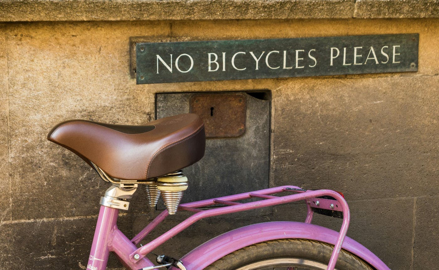 A purple bicycle leaning against a brown wall under a sign reading No Bicycles Please