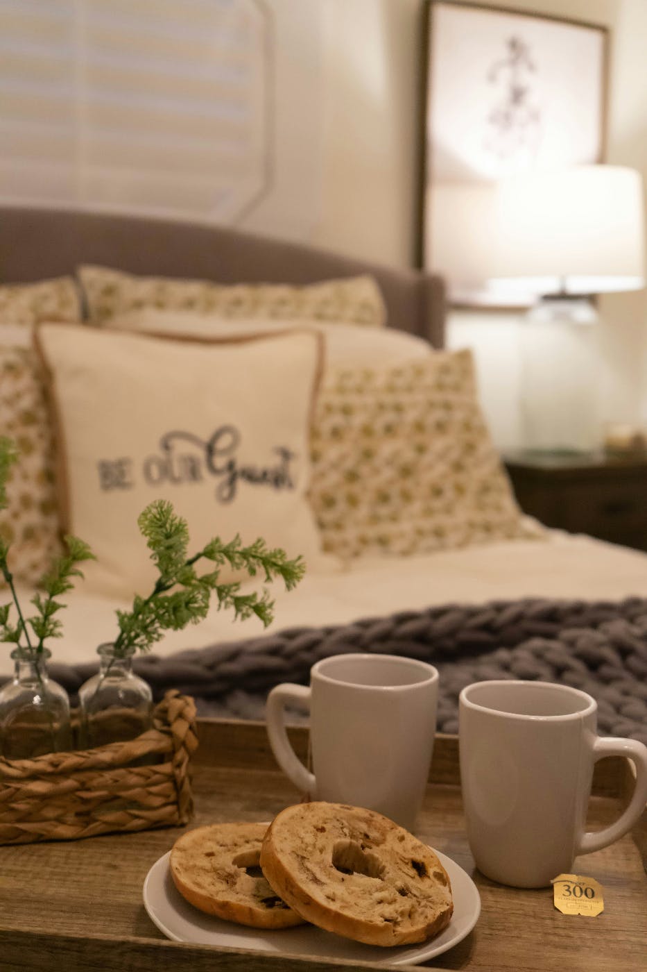 A guest room with coffee mugs and a bagel in the foreground and a pillow reading Be Our Guest on the bed