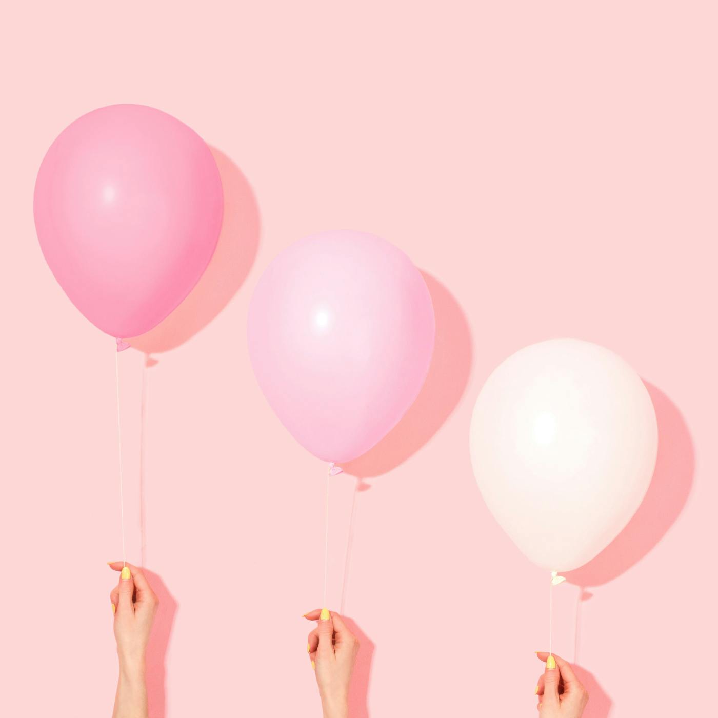 3 hands holding 3 balloons, pink, pale pink and white, against a light peach wall