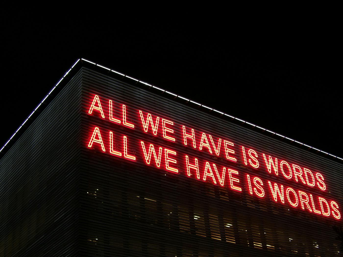 red neon sign on the side of a  building reading: All We Have Is Words, All We have Is Worlds