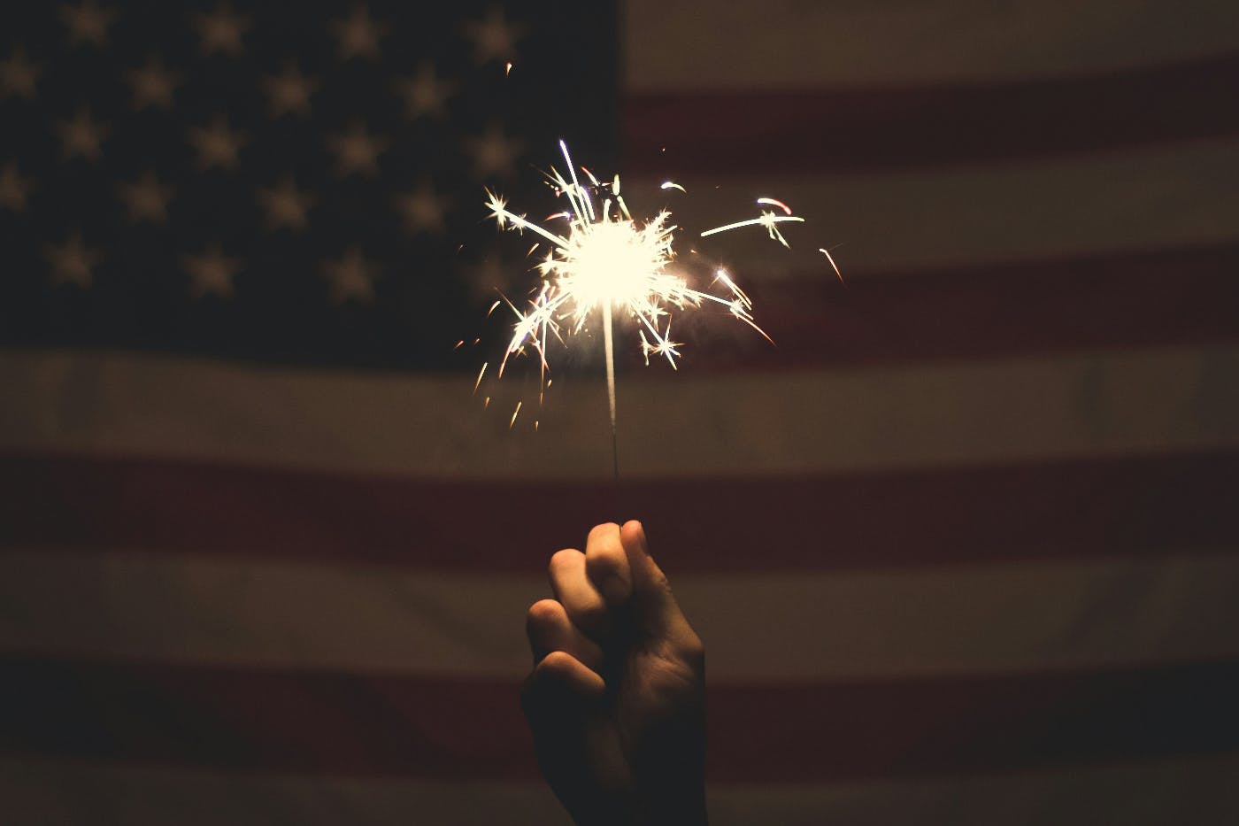 A hand holding a lit sparkler in front of an American Flag