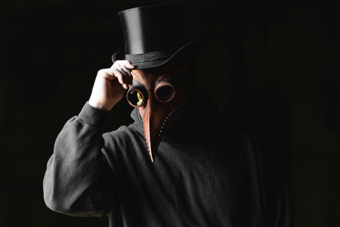 A man in black clothes, with a black top hat wearing a copper colored plague mask